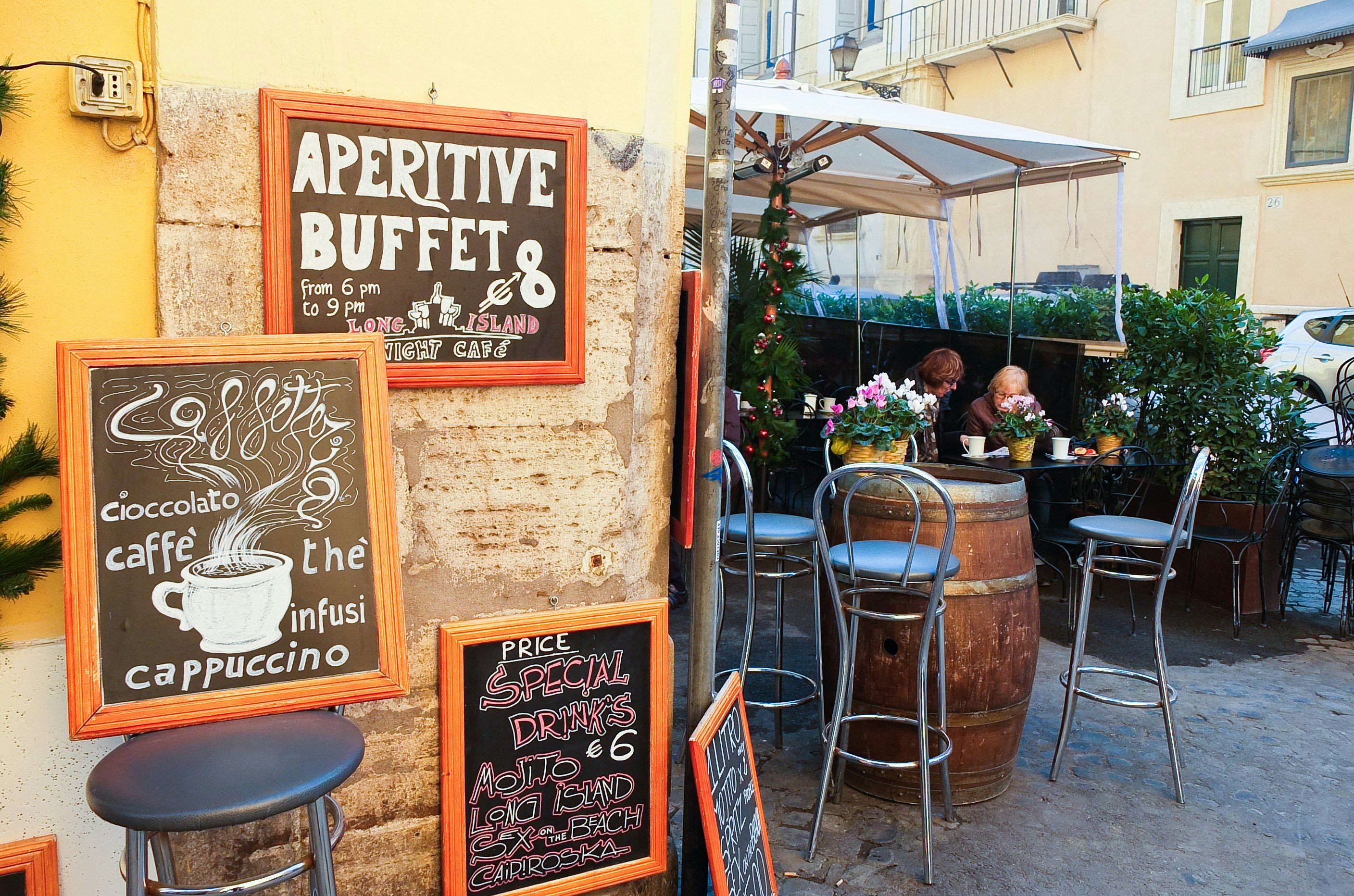 A bar with many blackboard signs advertising drinks, coffees, cappuccinos and cocktails with two older ladies drining coffee on an outside terrace.