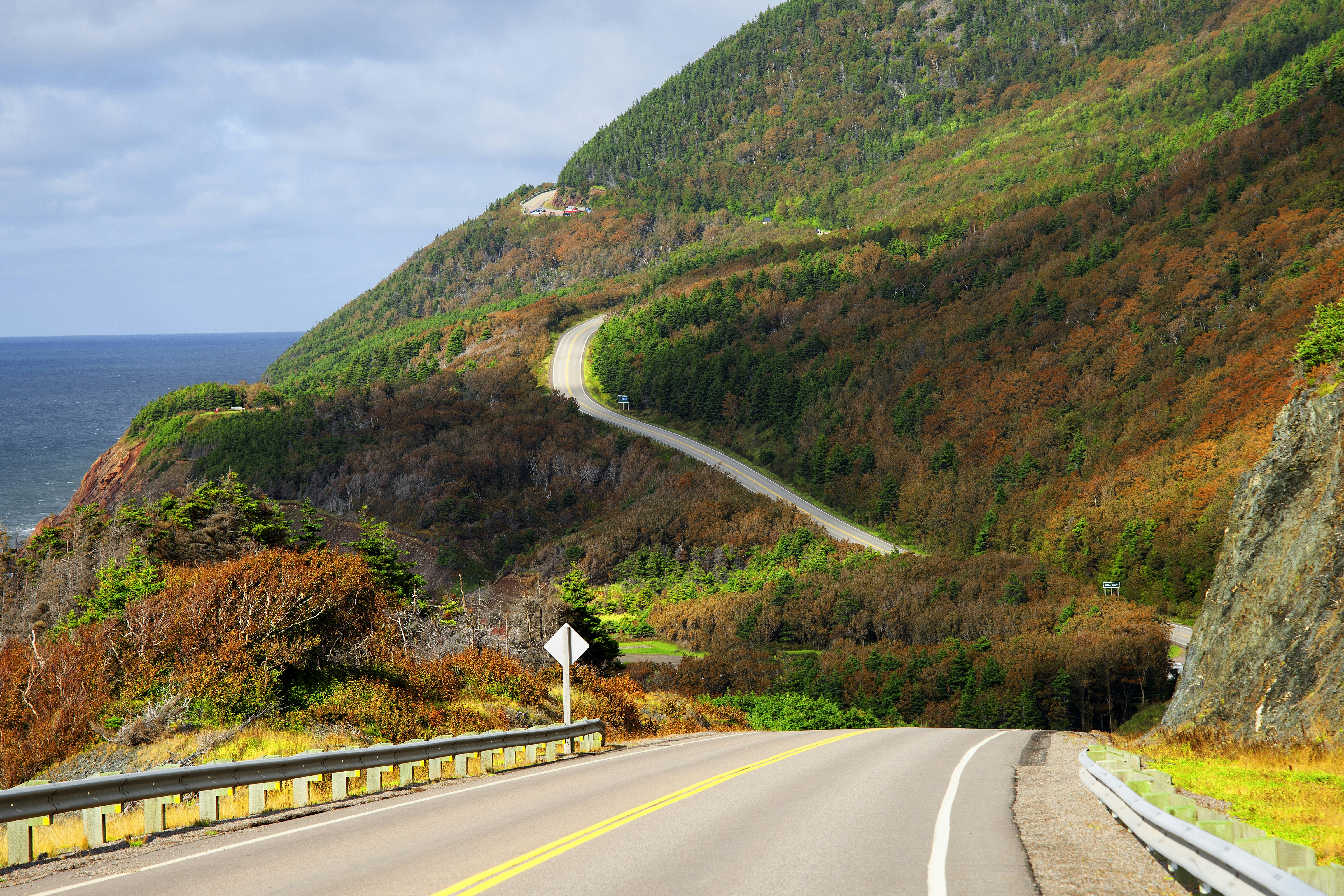 A road unspools with the sea on one side and dramatic fall foliage on the other, Cabot Trail, Cape Breton, Nova Scotia, Canada