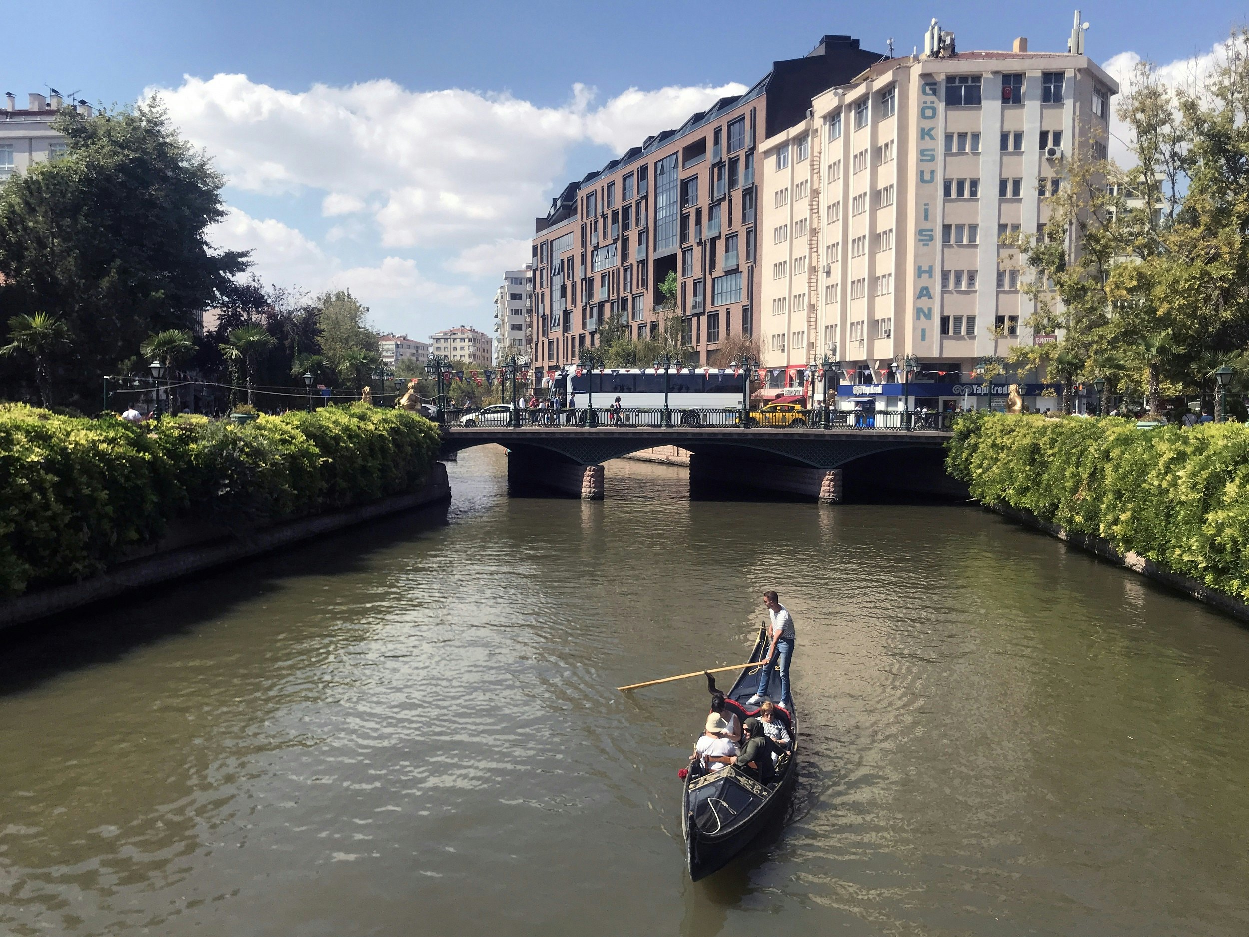 A gondola with three passengers travels down a river, which is lined with greenery