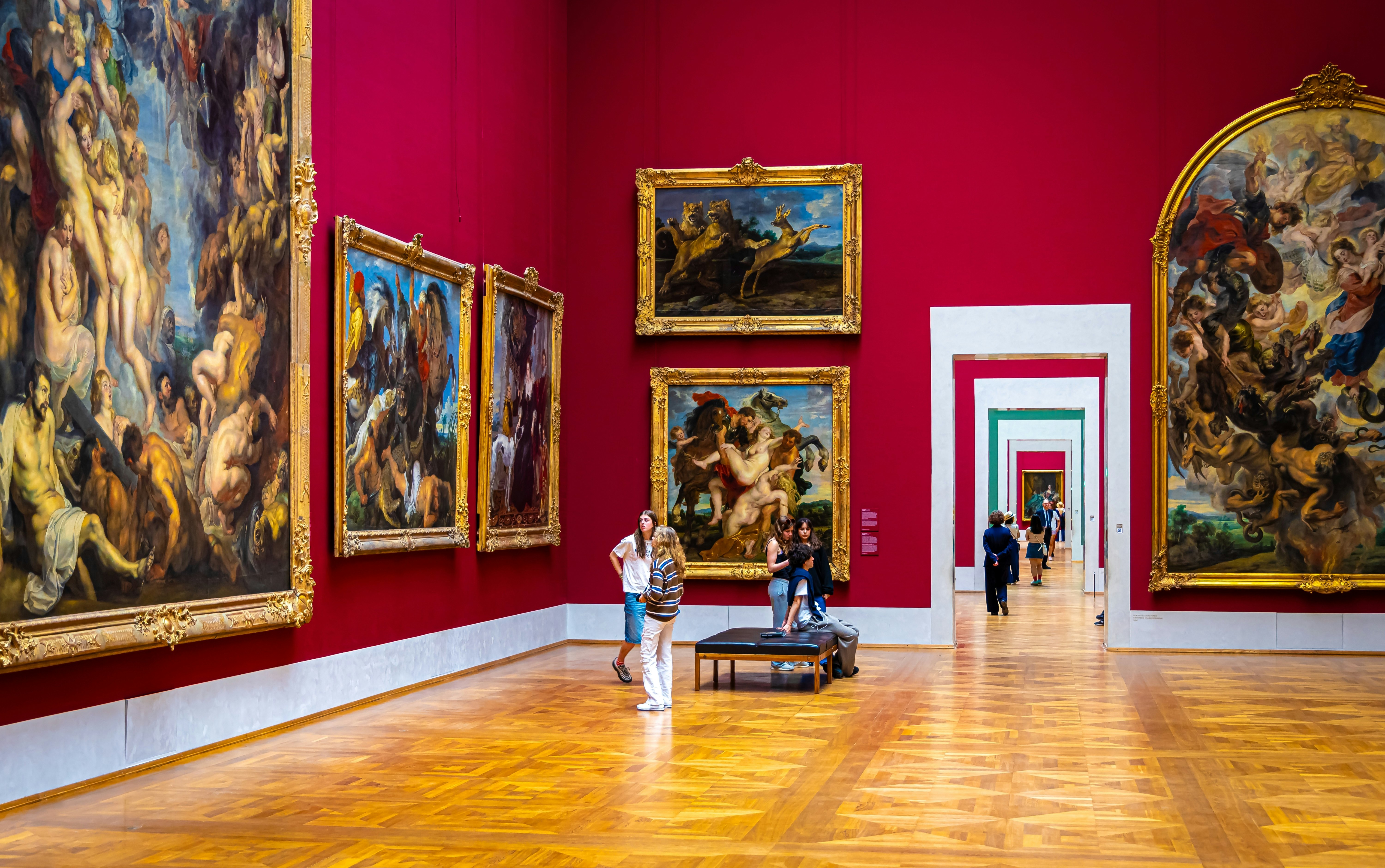 People look at large paintings in a museum gallery with red walls. Other galleries can be seen through a doorway, en enfilade.