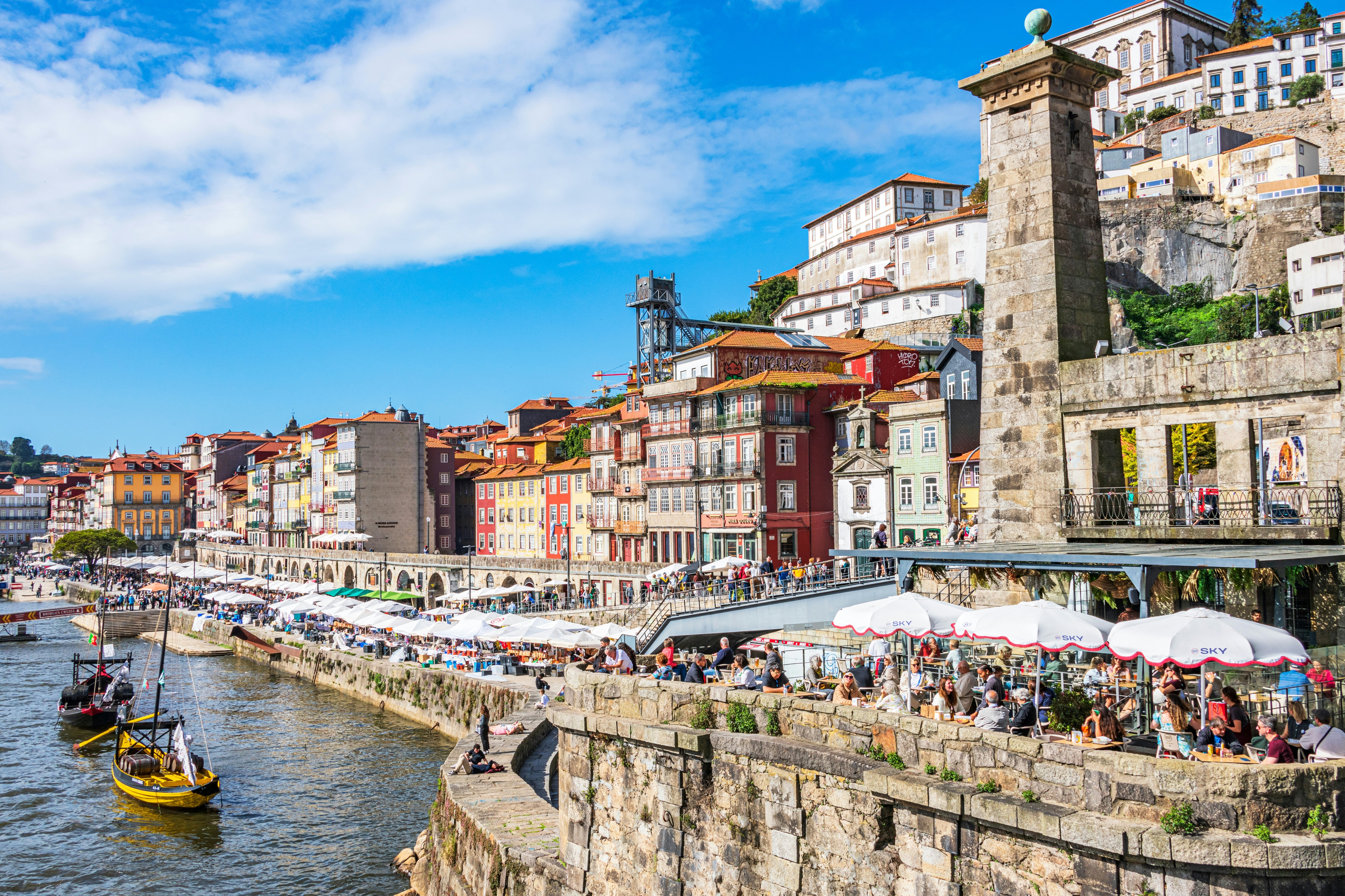 View of a riverfront with cafe umbrellas lining a cobblestone wall and colorful buildings to the right.