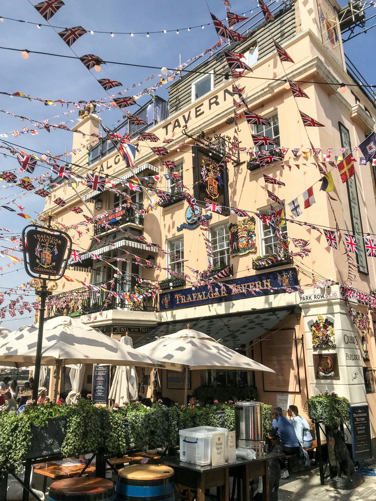 People sit in the shade of a terrace that surrounds a pub called the Trafalgar Tavern, with flag-themed bunting flying all around.